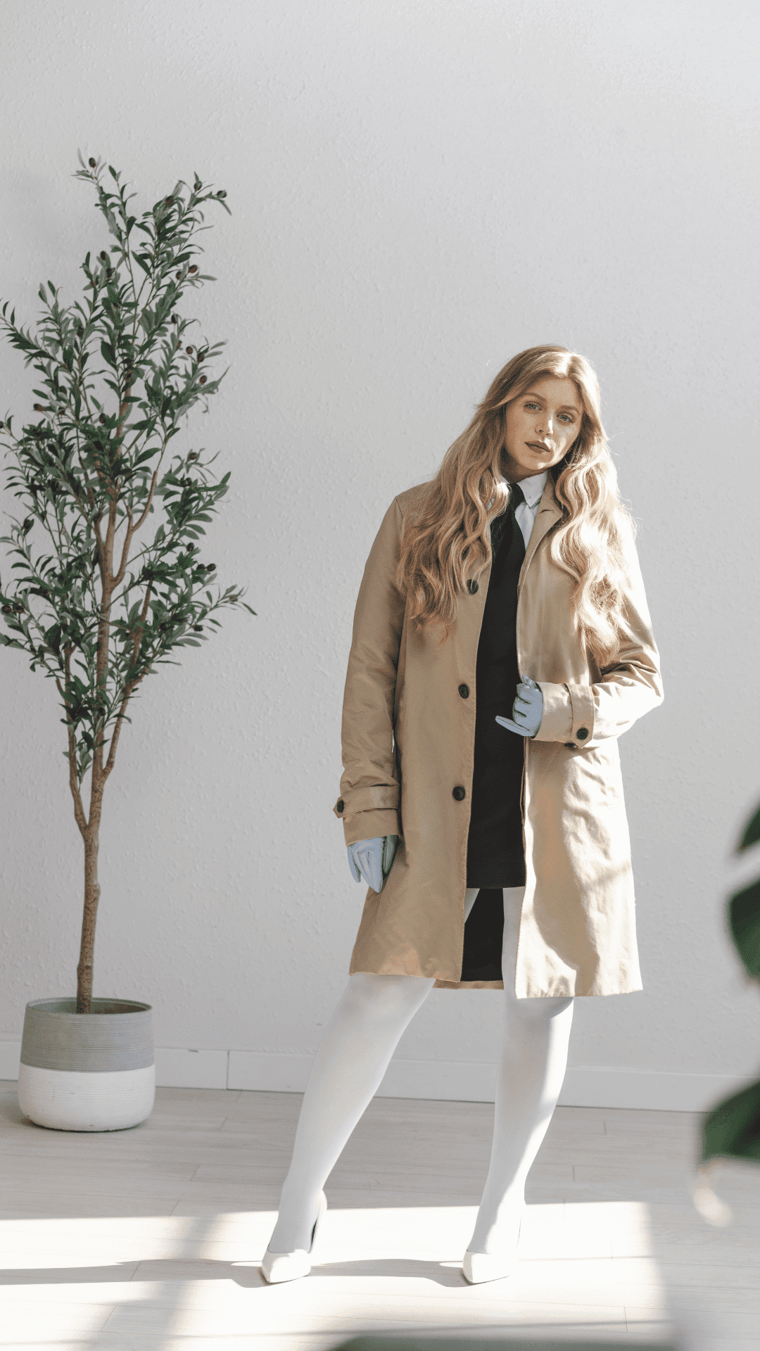 Blonde woman in beige coat and gloves, standing indoors next to a potted plant.