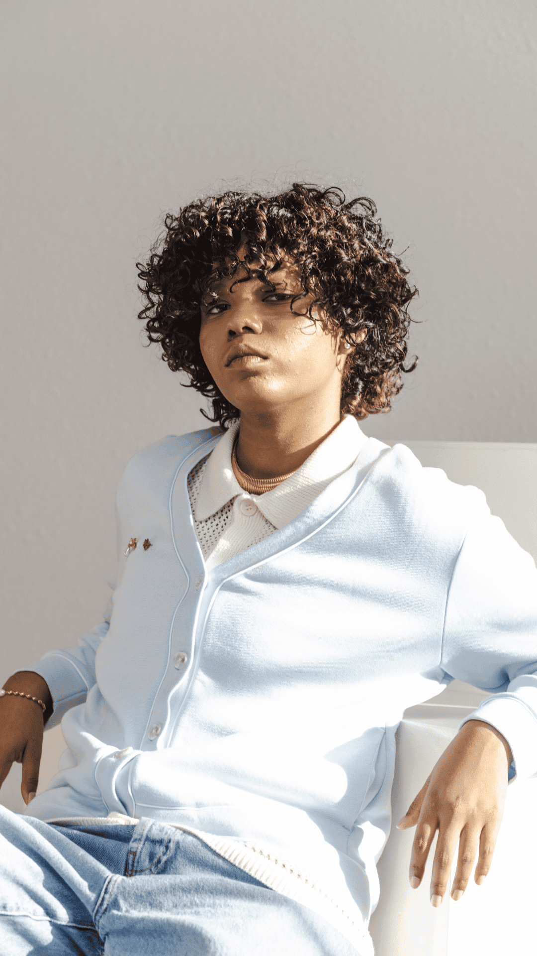 A person with curly hair in a light blue cardigan, sitting against a plain backdrop.