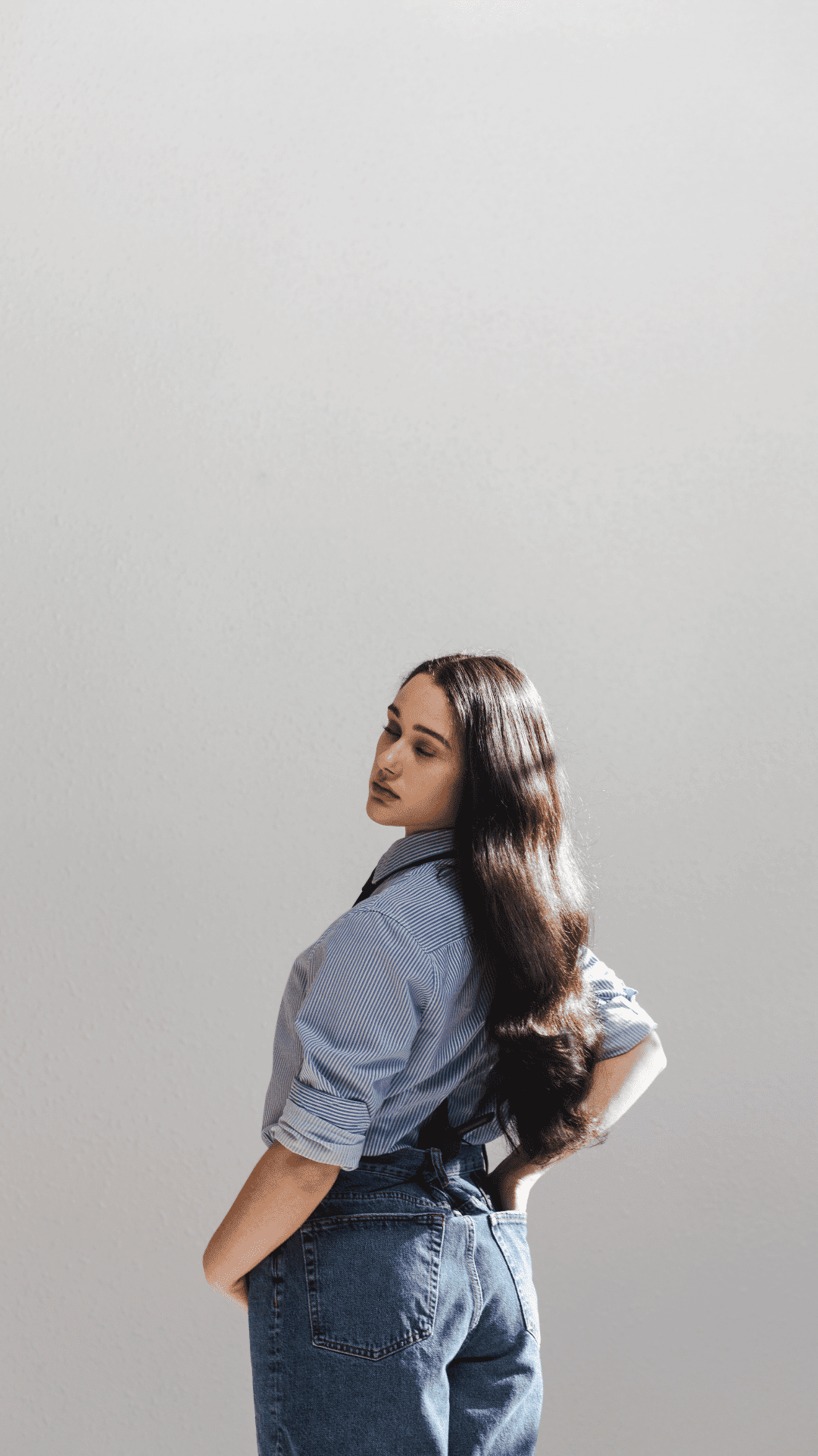 Woman in blue shirt with long hair stands against a light background, looking to the side.