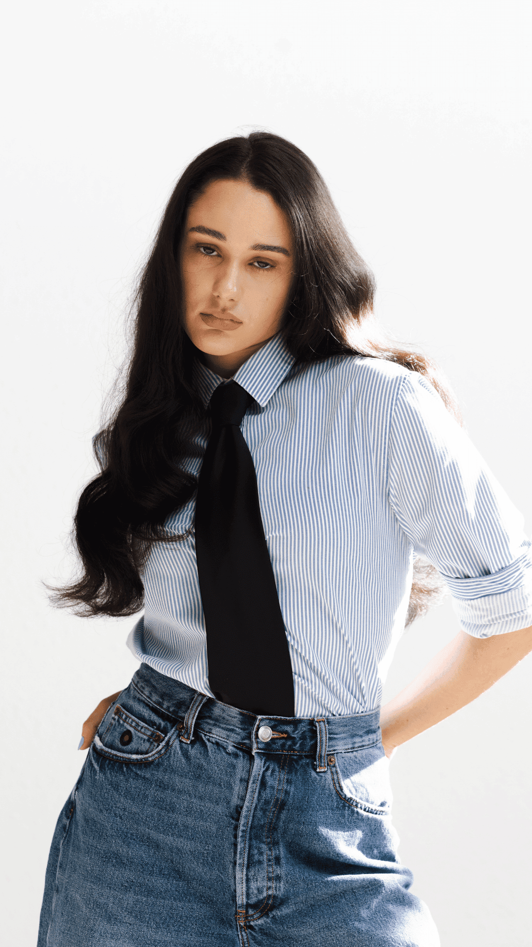 Woman in striped shirt and tie with hands in jean pockets.