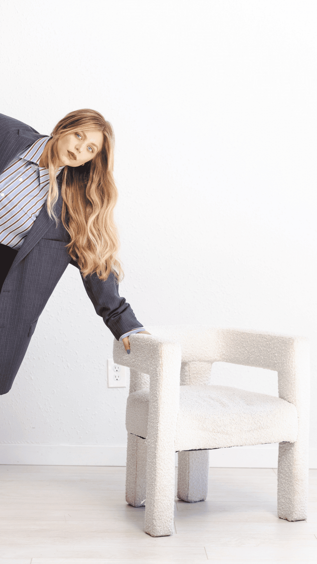 Woman in gray blazer leaning on a textured white chair in a minimalist room.