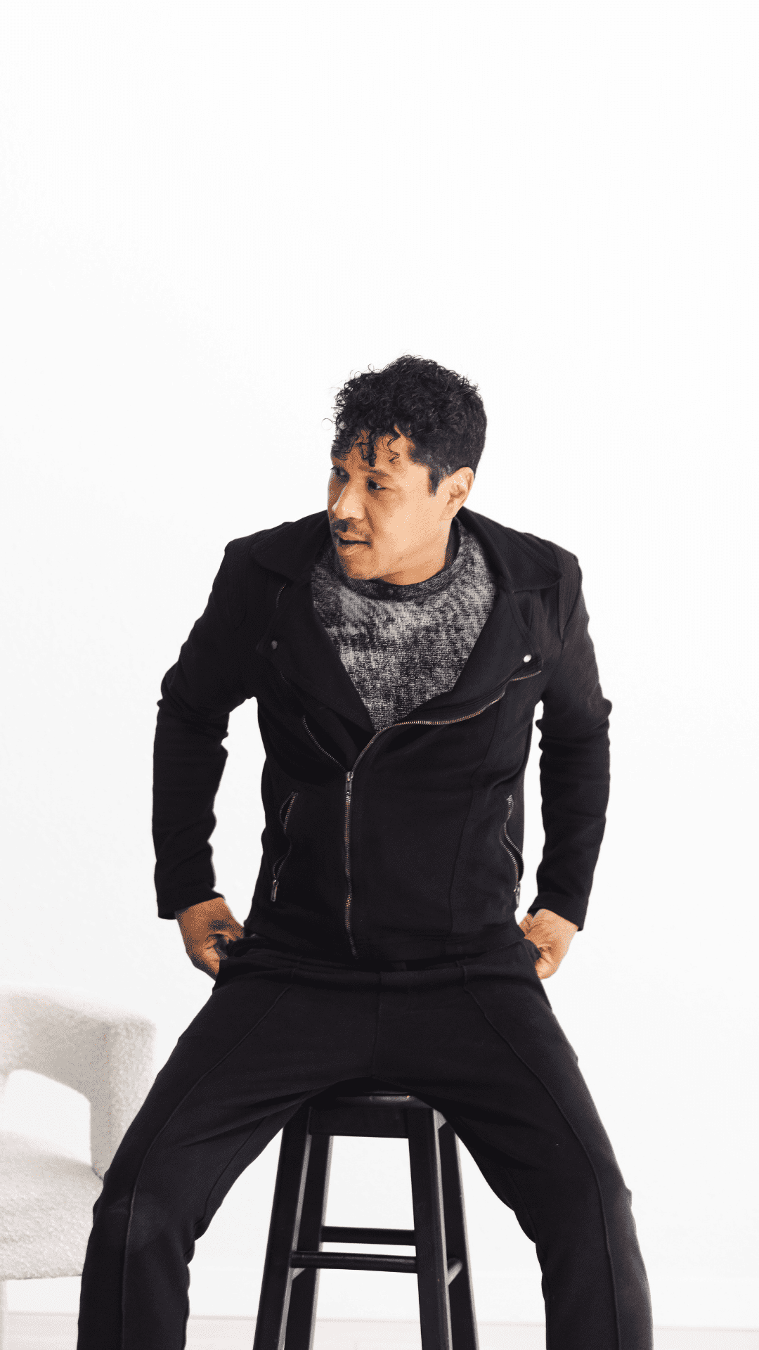 Man in black jacket sitting on a stool against a white background.