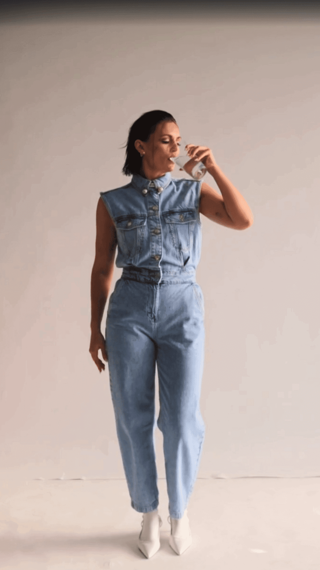 Woman in denim jumpsuit drinks from a cup, standing with relaxed posture.