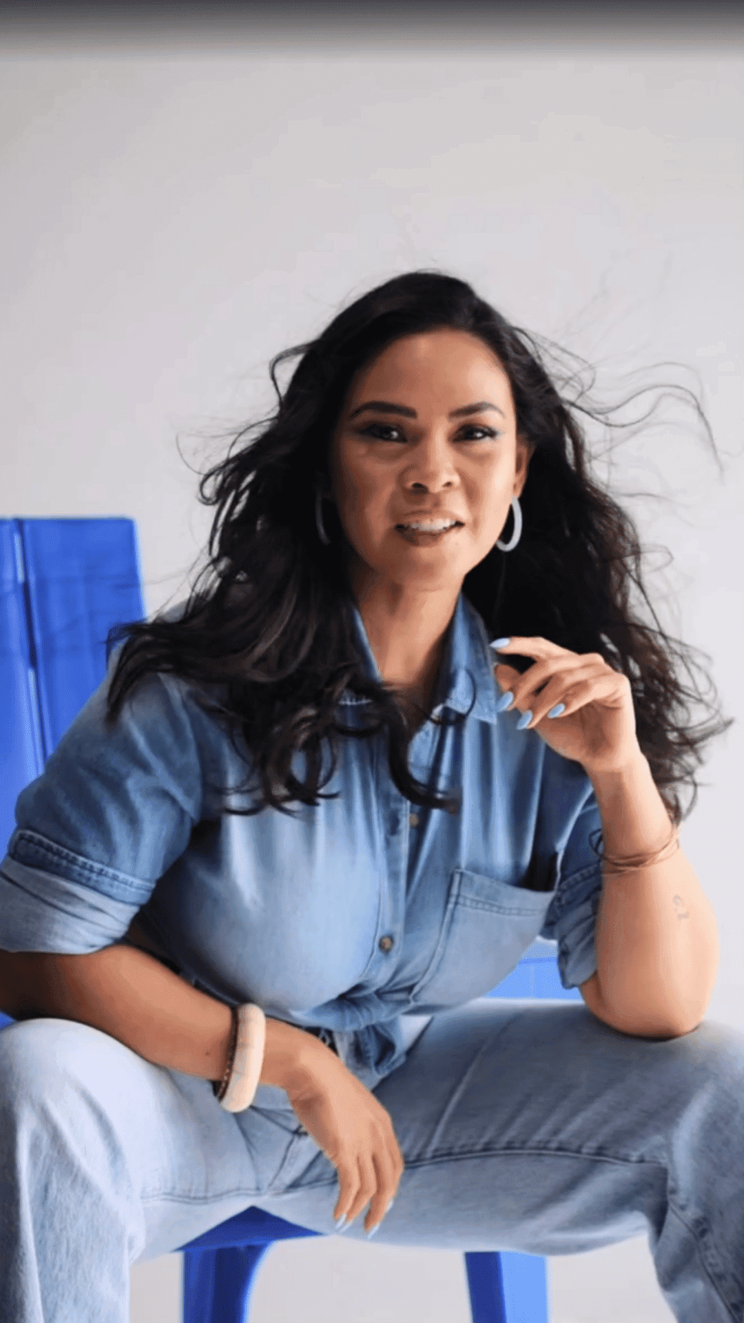 Woman seated on a blue chair, wearing a denim outfit and hoop earrings, smiling softly.