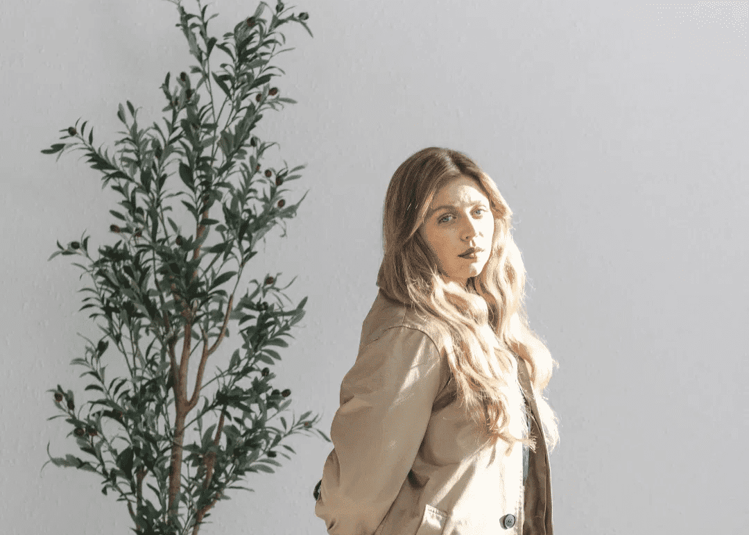 A woman in a beige coat stands elegantly by a potted plant under soft lighting.