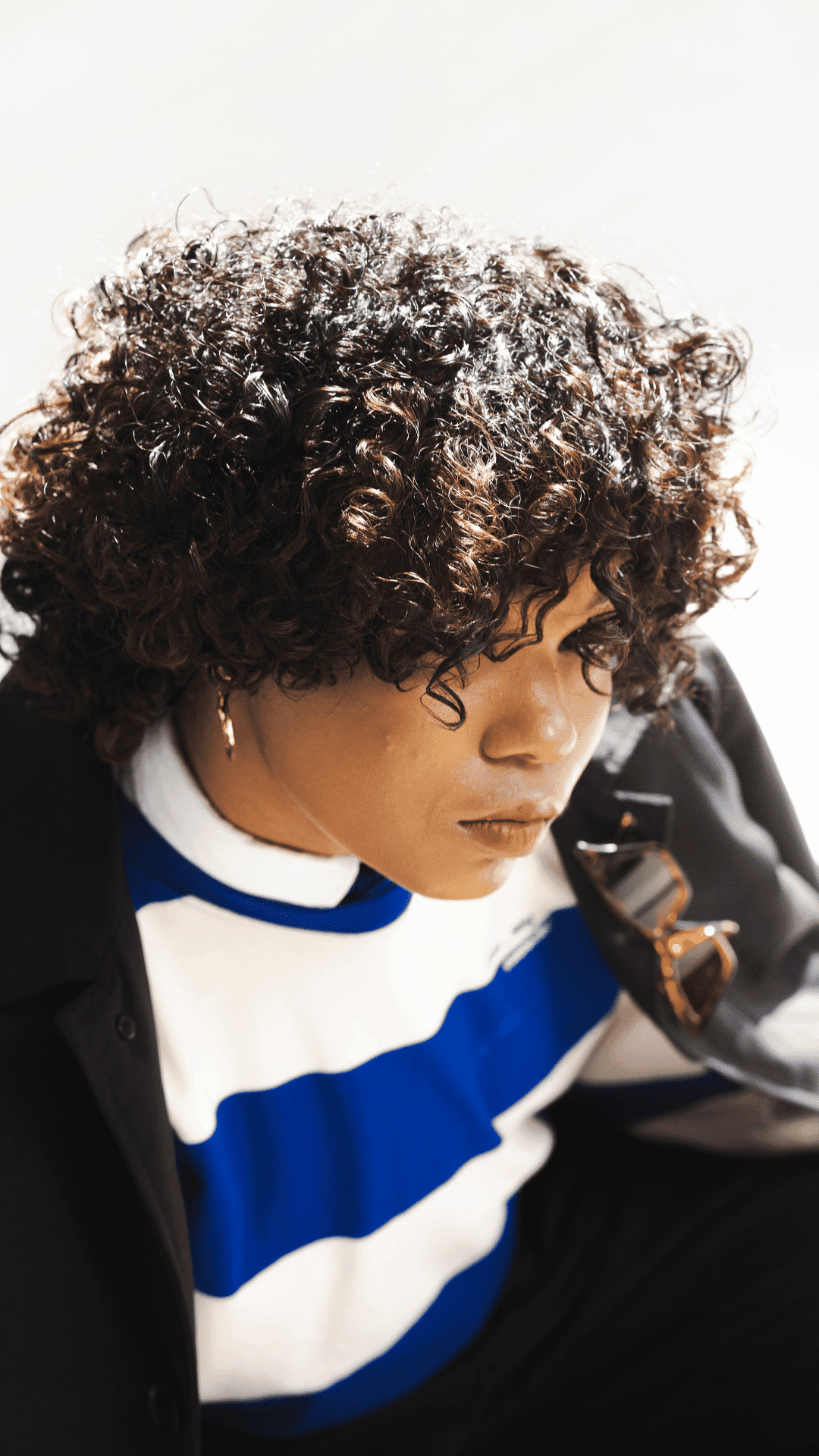 Curly-haired person in blue and white striped shirt, wearing a coat and holding sunglasses.
