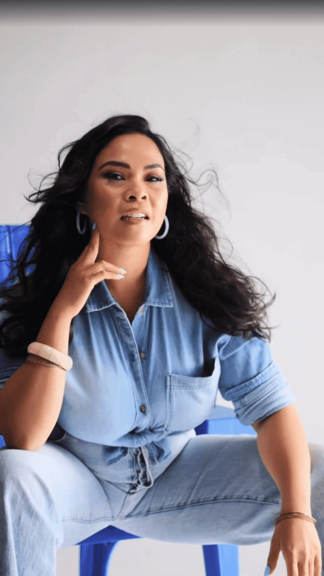 Jeans-clad woman seated on a blue chair, long wavy hair, with a confident, relaxed pose.